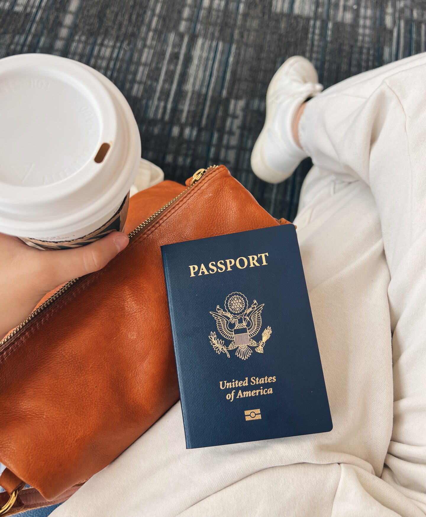 Girl sitting crossed-legged in an airport with coffee cup and passport.